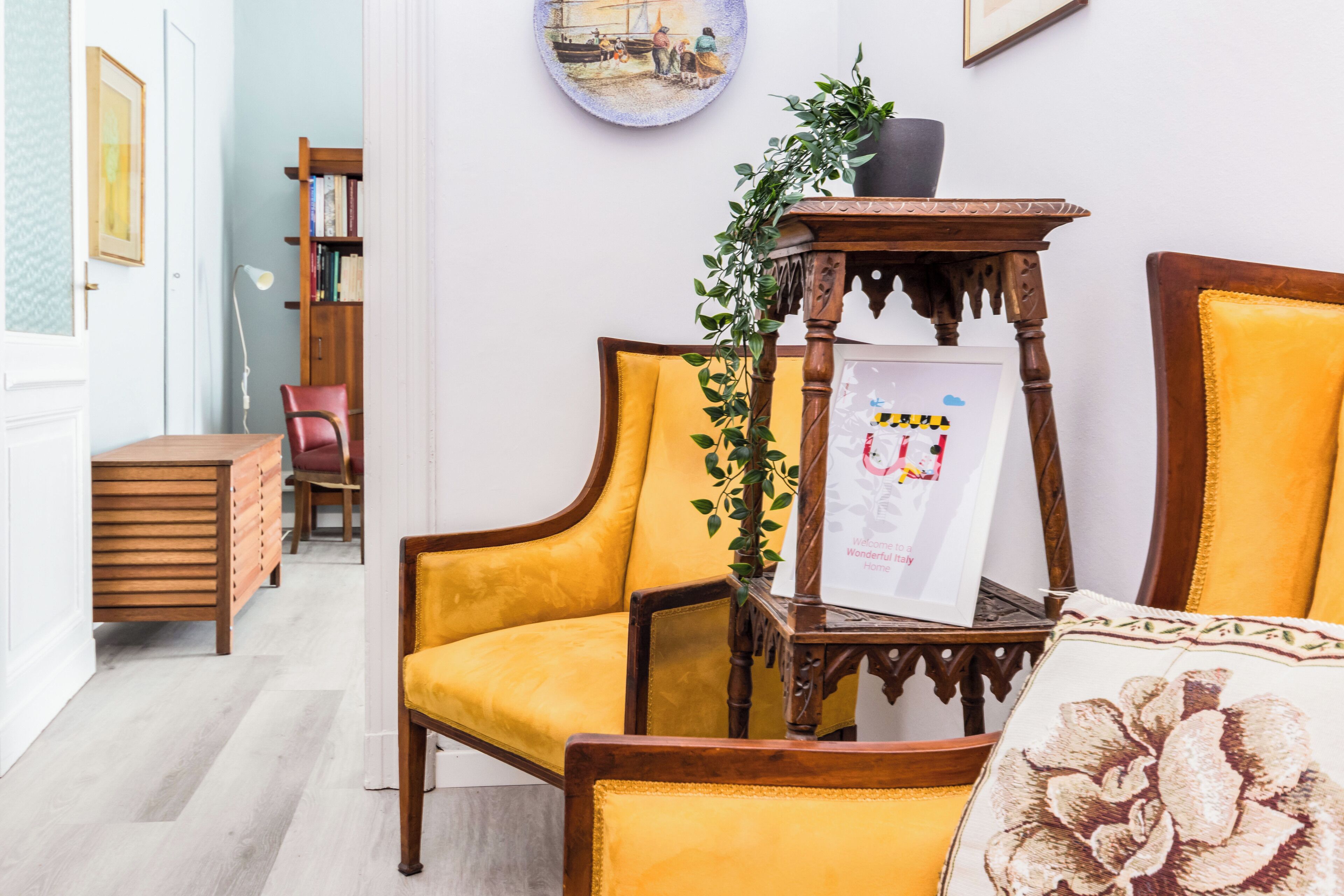 A room with a wooden chair, a small table with a plant, and a framed welcome sign.
