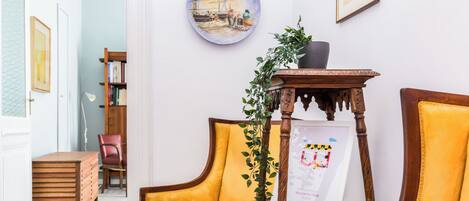 Una habitación con una silla de madera, una mesita con una planta y un cartel de bienvenida enmarcado.