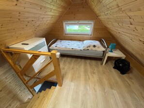 A small, wooden-paneled room with a bunk bed, a desk with a computer, a window, and a fan.