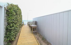 A narrow wooden pathway leading to a beach area with a bench and a table, surrounded by a hedge and a white wall. - Sun Realty cozy 4BR oceanside Duck cottage just 300' to the beach (Duck)