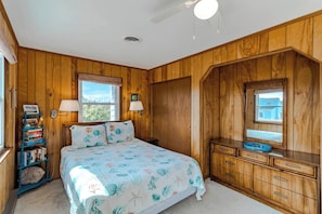 A bedroom with a bed, a dresser, a lamp, a bookshelf, and a window. - Sun Realty cozy 4BR oceanside Duck cottage just 300' to the beach (Duck)