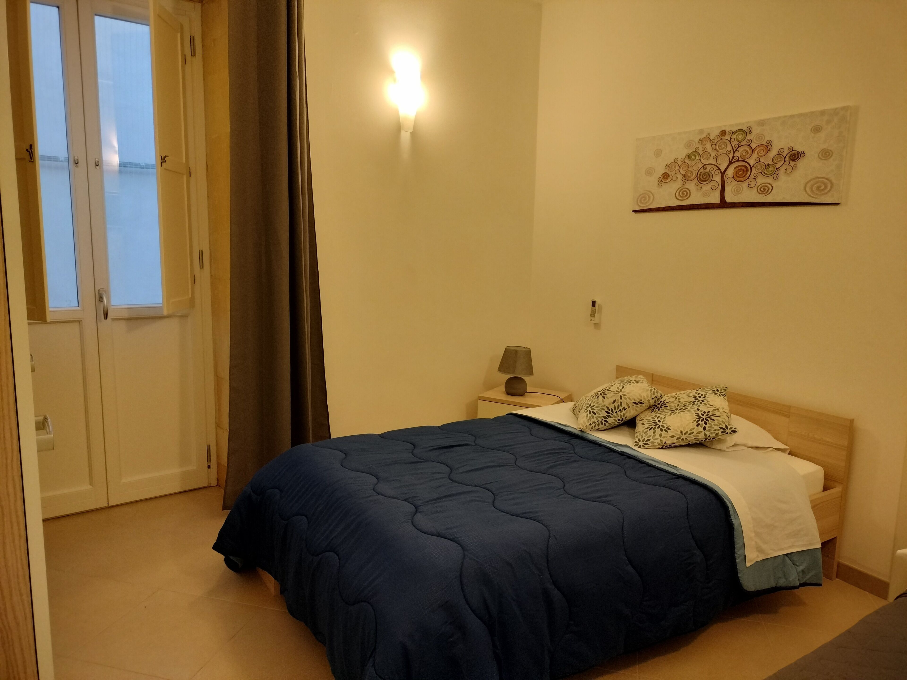 Une chambre à coucher avec un lit, une table de chevet, une lampe, un tableau et une porte avec des rideaux.