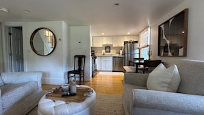 A modern living room with a grey sofa, a wooden coffee table, and a dining area with a table and chairs. A kitchen with white cabinets and a refrigerator is visible in the background. - Ptown Paradise Farmhouse in the perfect, central location! (Provincetown)