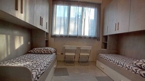 Quarto compacto com duas camas de solteiro, uma mesa pequena e uma janela com cortinas.