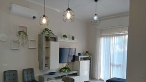 Uma área de jantar moderna com uma mesa retangular posta para quatro pessoas, rodeada por cadeiras. Há três luminárias pendentes sobre a mesa e uma janela grande com persianas à direita.