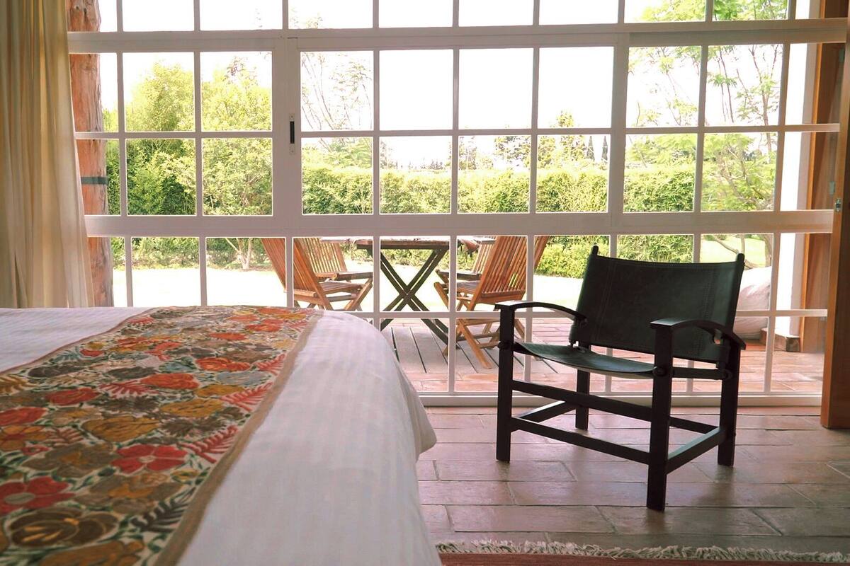 A bedroom with a large bed, a chair, and a view through a window to a patio area.