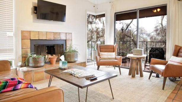 A modern living room with a fireplace, comfortable seating, a flat-screen TV, and a large window offering a view of trees. - Downtown Treetop Hideaway- SXSW, 6th St, UT Campus

 (Austin)