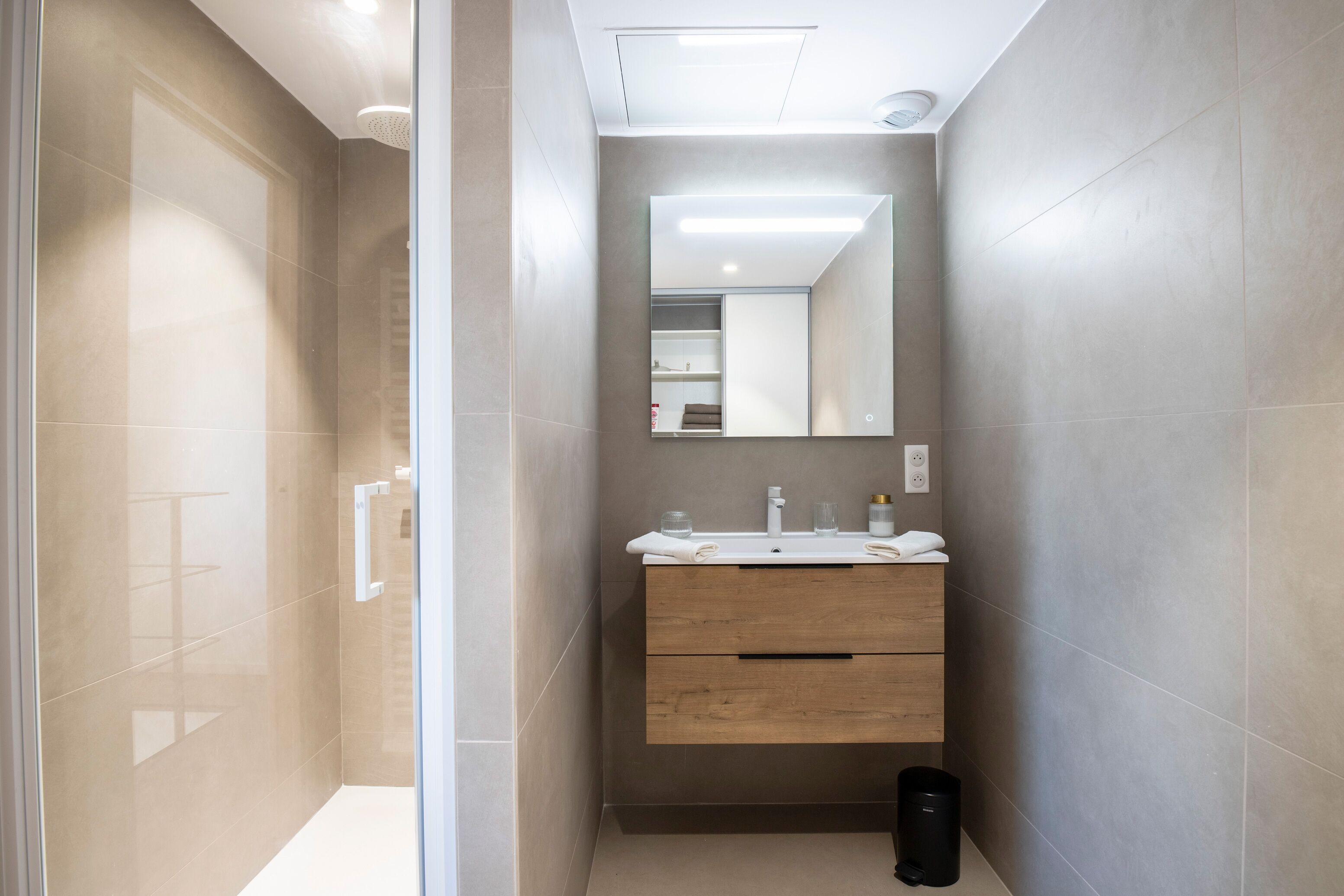 A modern bathroom with a glass shower enclosure, a wooden vanity with a mirror, and a black trash bin.