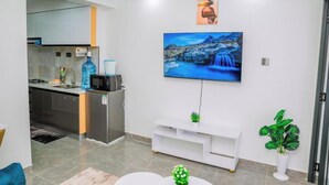 A modern living room with a flat-screen TV mounted on the wall, a white entertainment unit, and a kitchen area visible through an open door. - Lulu Homes at Gatundu Heights (Nairobi)