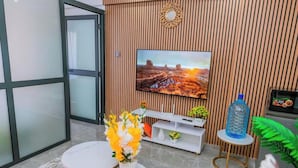 A modern living room with a flat-screen TV mounted on a wooden wall, a white entertainment unit, and two round tables with floral centerpieces. - Lulu Homes at Gatundu Heights (Nairobi)
