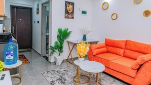 A modern living room with a bright orange sofa, a white coffee table, and a small white table. There is a large water bottle on a side table, a potted plant, and a framed artwork on the wall. - Lulu Homes at Gatundu Heights (Nairobi)