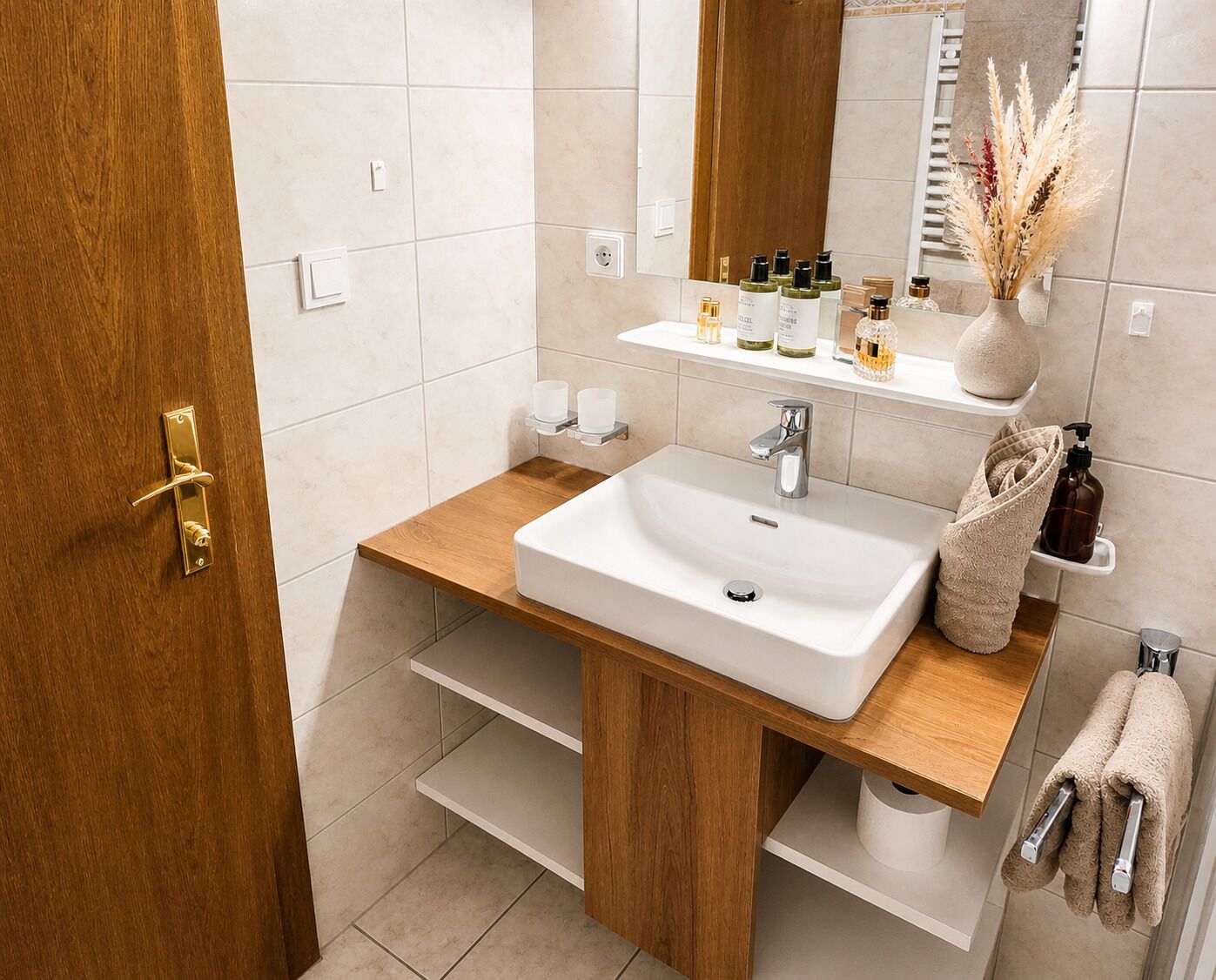 A bathroom with a wooden vanity, white sink, and a mirror. There are toiletries on the vanity and a towel rack with towels.
