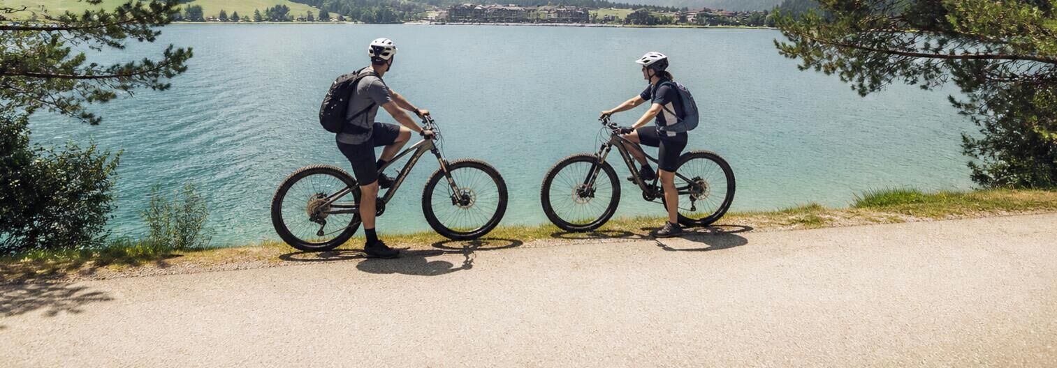 Deux cyclistes sur une piste cyclable asphaltée, au bord d’un lac, avec une vue dégagée sur l’eau et les bâtiments situés au loin.