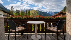 Een houten terras met een ronde tafel en twee rieten stoelen, met uitzicht op een bergachtig landschap en rode bloemen op de voorgrond.