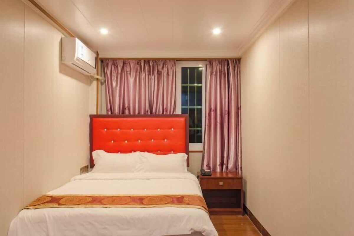 Een hotelkamer met een rood hoofdeinde, een bed met witte beddengoed, een nachtkastje, een raam met gordijnen en een airconditioner.