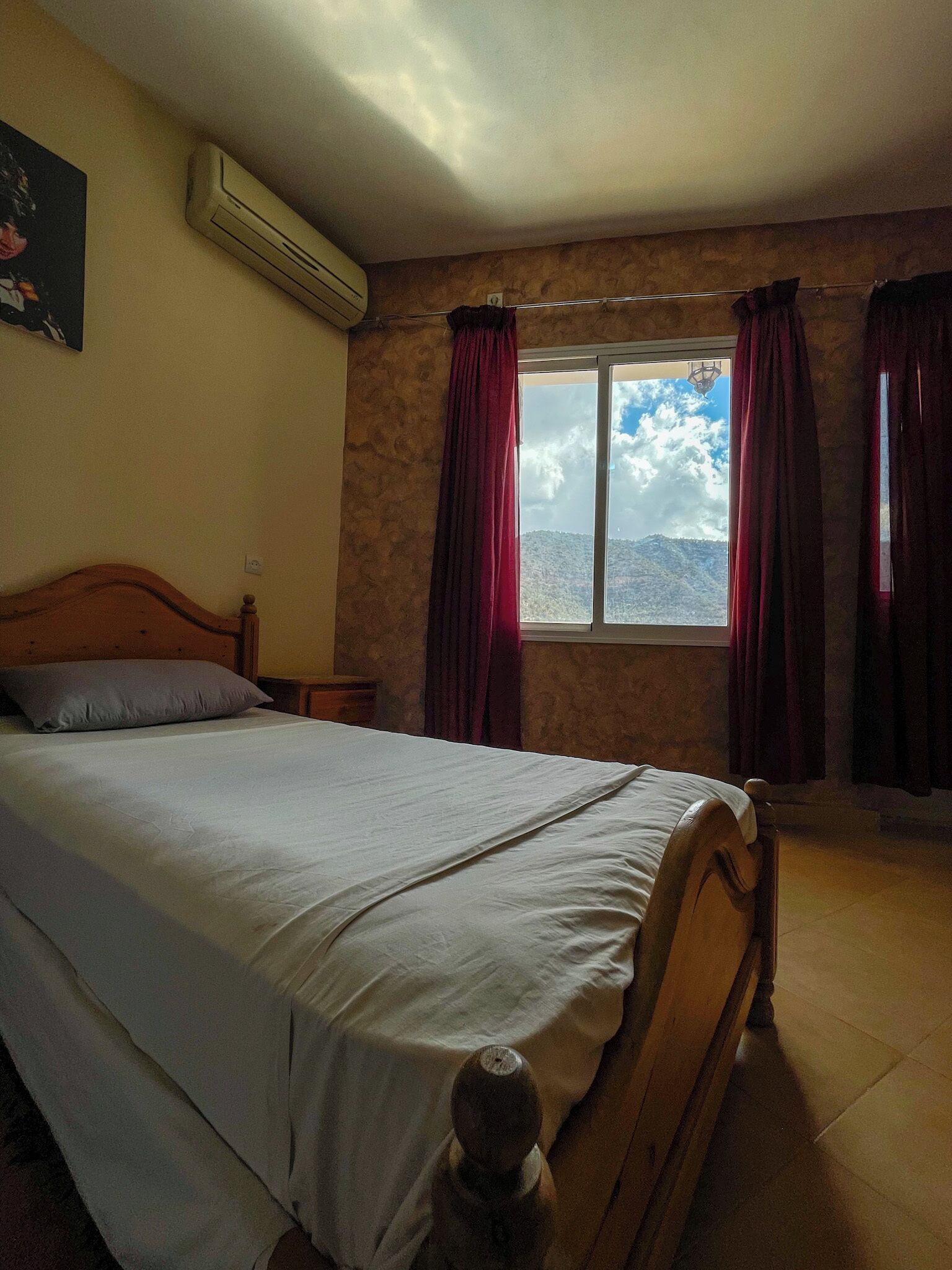 A bedroom with a bed, a window with curtains, a wall-mounted air conditioner, and a painting on the wall.