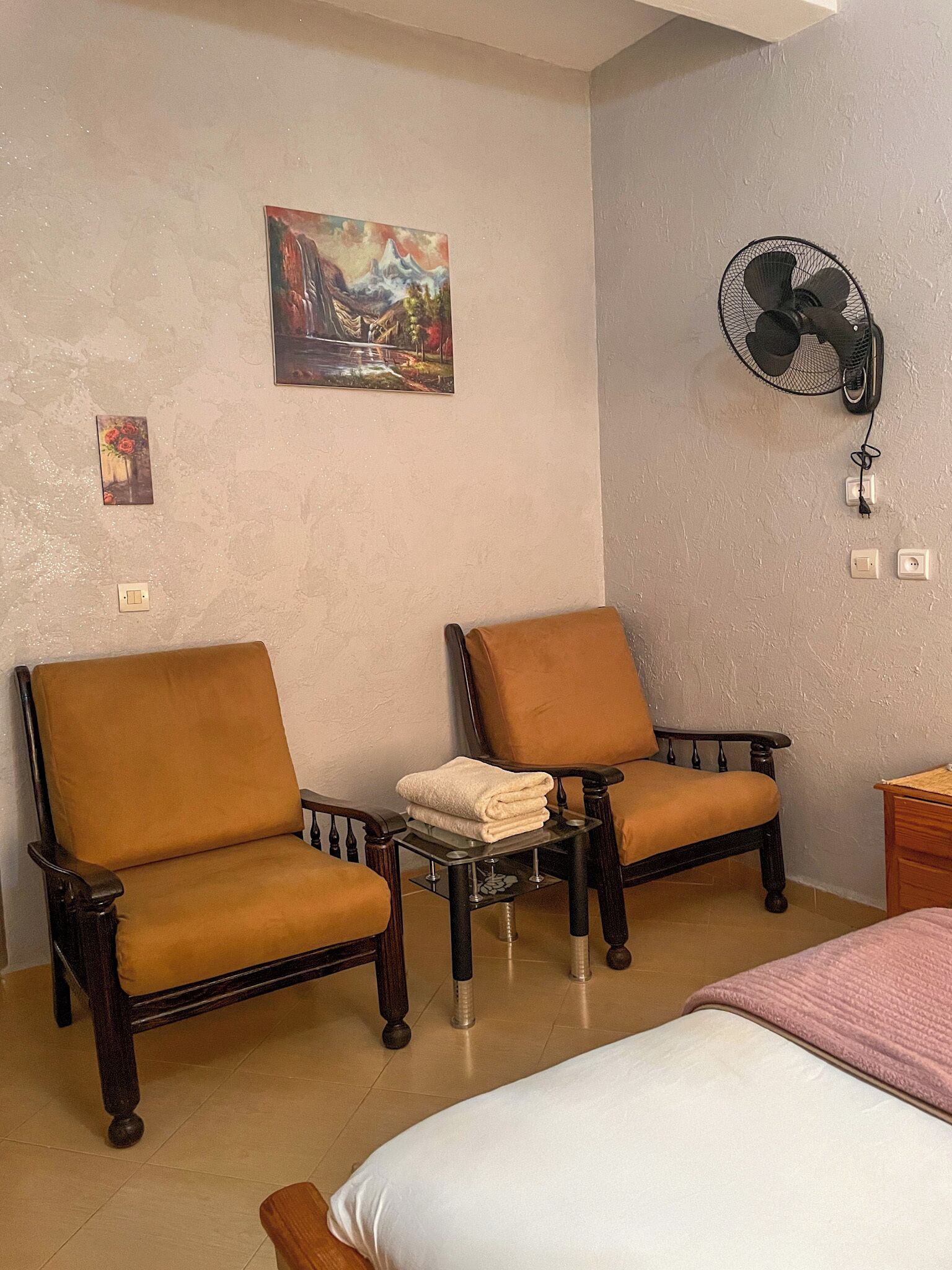 A hotel room with two brown armchairs, a bed with a white mattress, a small table with a folded towel, a wall-mounted fan, and a painting above the armchairs.