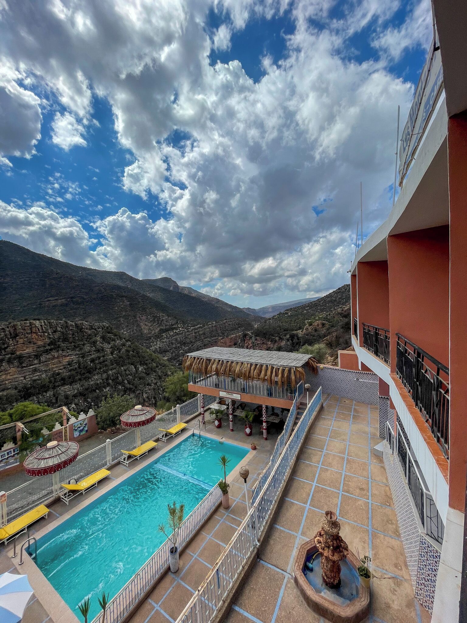 A rooftop pool area with a clear view of the surrounding mountains, a pool with yellow loungers, and a building with red and white walls.