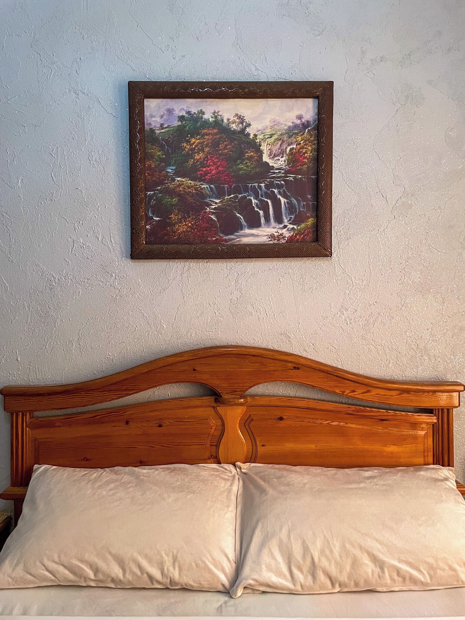 Ein Bett mit einem hölzernen Kopfteil und zwei Kissen, ein gerahmtes Gemälde eines Wasserfalls an der Wand darüber.