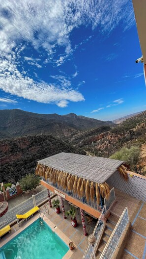 Un espace piscine offrant une vue sur les montagnes, un coin salon abrité et un panneau indiquant l’emplacement d’un restaurant.