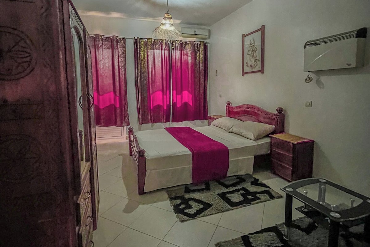 Une chambre à coucher comprenant un lit, une armoire, un climatiseur, un ventilateur de plafond et une petite table.