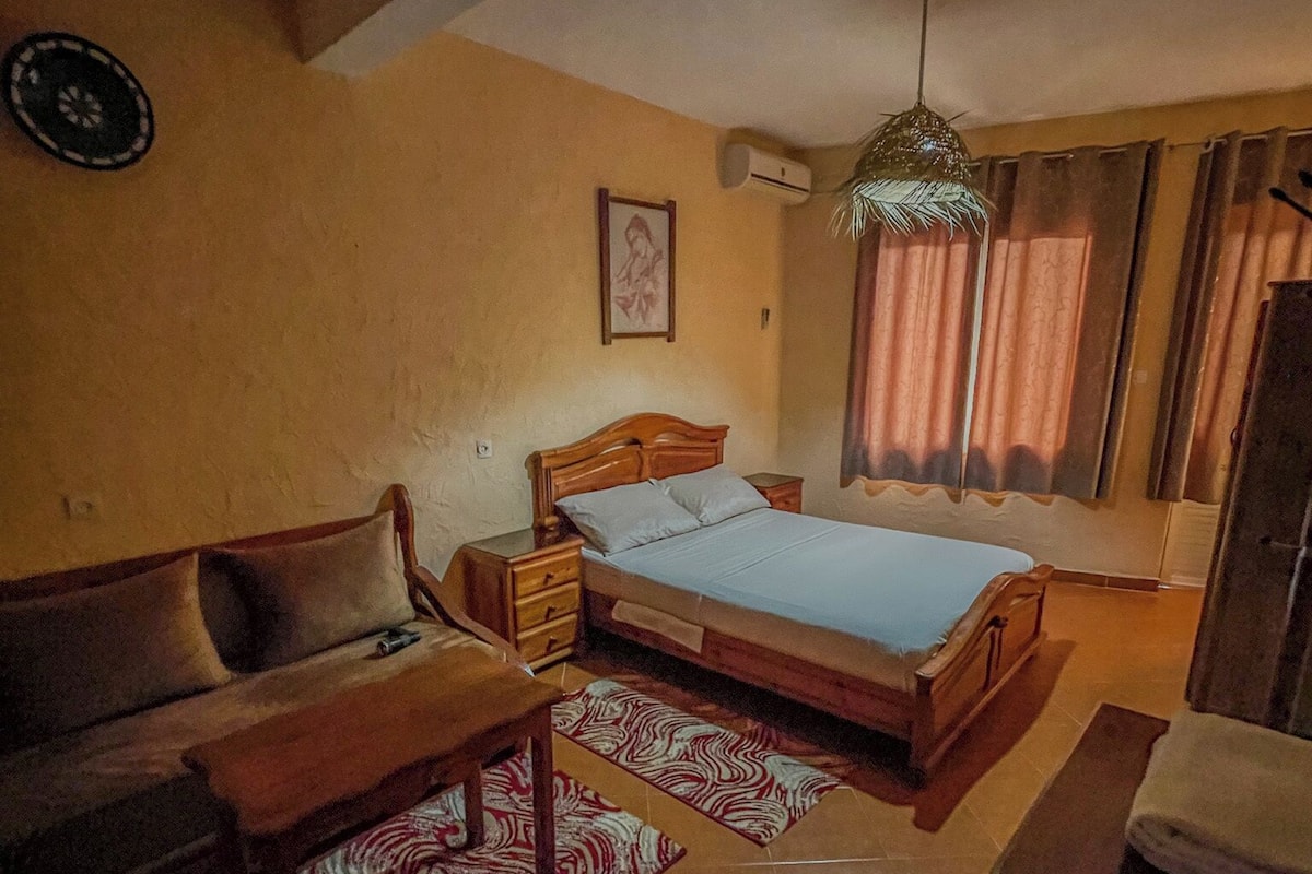 Une chambre à coucher avec un lit, une table de chevet en bois, un bureau et une chaise. Il y a une fenêtre avec des rideaux et un appareil de climatisation fixé au mur.