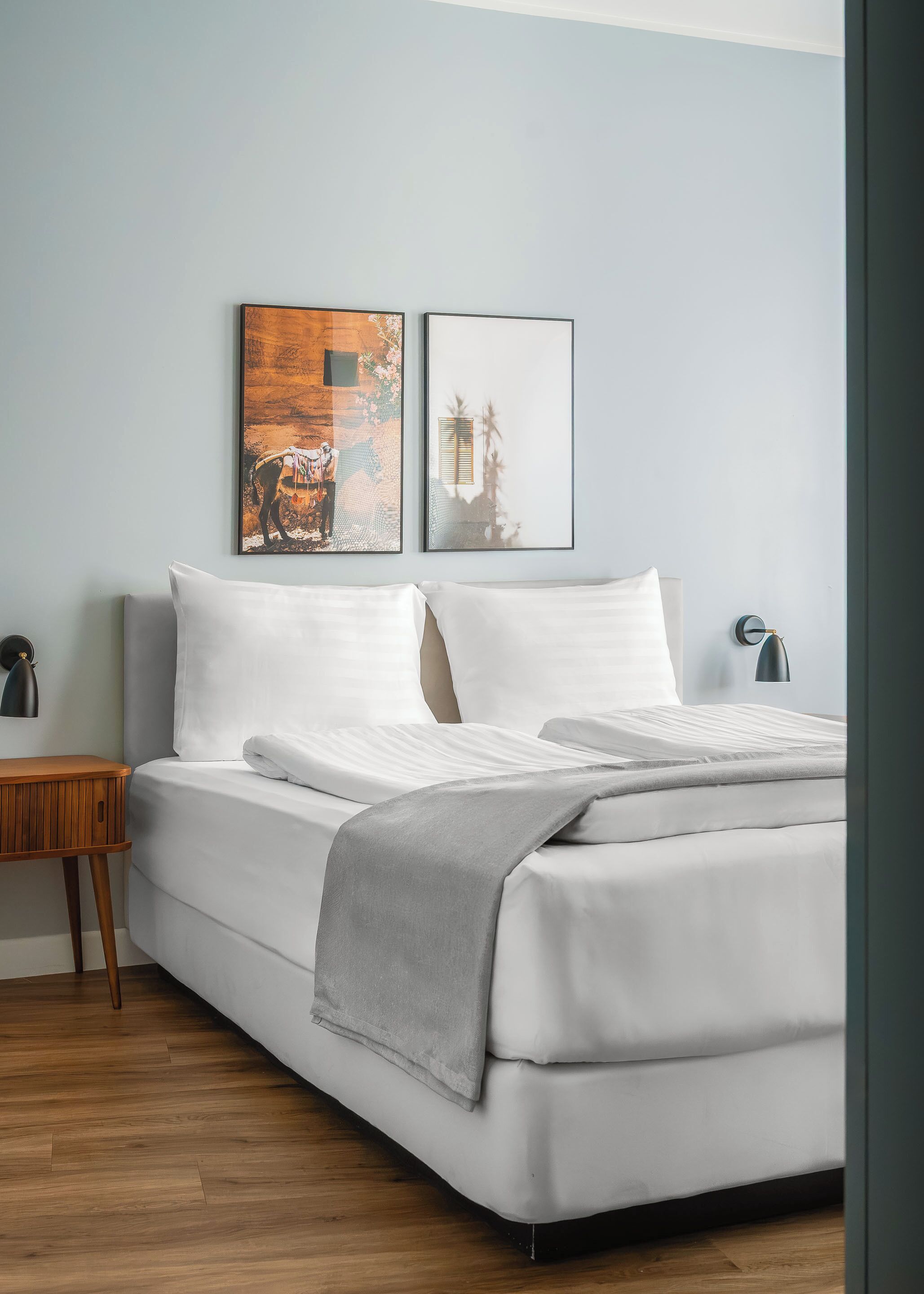 A neatly made bed with white linens and a gray throw blanket, a wooden nightstand with a lamp, and two framed pictures on the wall.