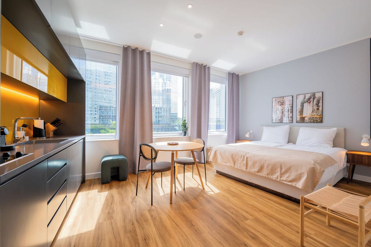 Una habitación de hotel moderna con una cama grande, una mesa de comedor con sillas, una zona de cocina y ventanas amplias con cortinas.