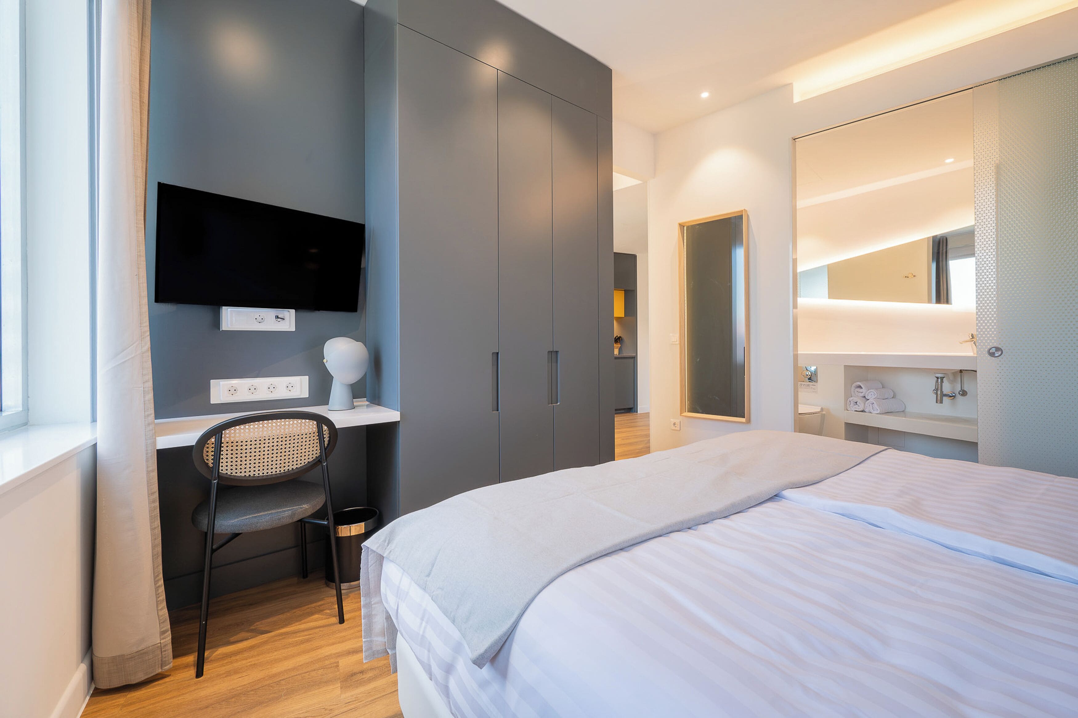 Un dormitorio moderno con cama, escritorio, silla, televisor y un espejo grande.