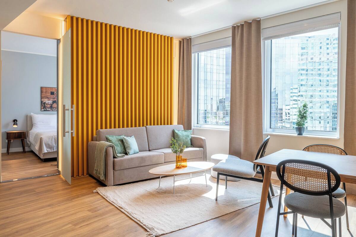 Un salón moderno con sofá, mesa de centro y comedor. Un ventanal grande ofrece vista a la ciudad.