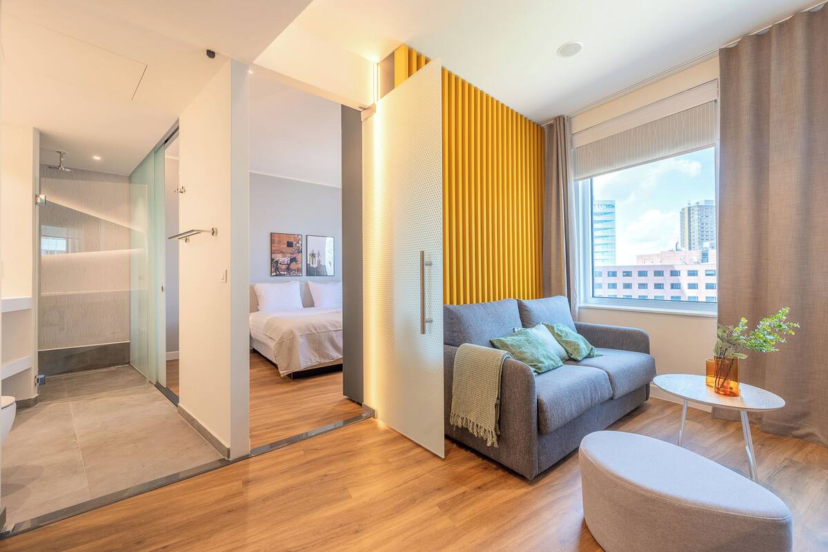 Habitación de hotel moderna con piso de madera, un sofá gris con una manta, una pequeña mesa blanca y una ventana con cortinas.