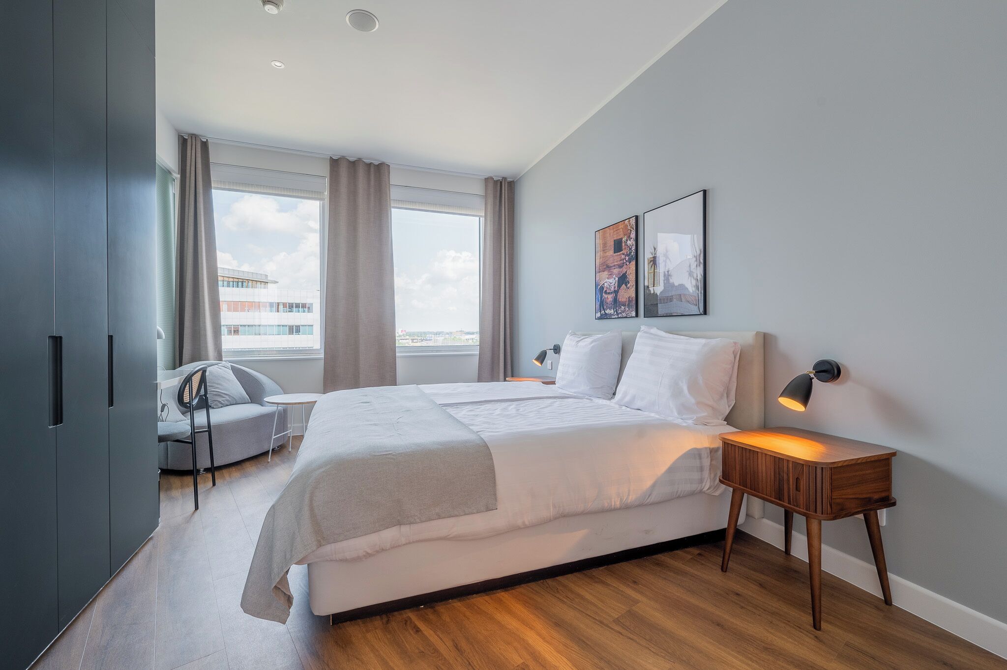 Een moderne hotelkamer met een groot bed, een nachtkastje met lamp, een stoel en uitzicht op de stad door grote ramen.