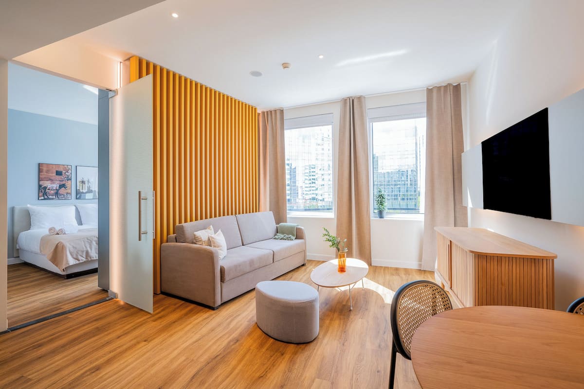 Habitación de hotel moderna con sofá, reposapiés, mesa redonda y televisor montado en la pared.