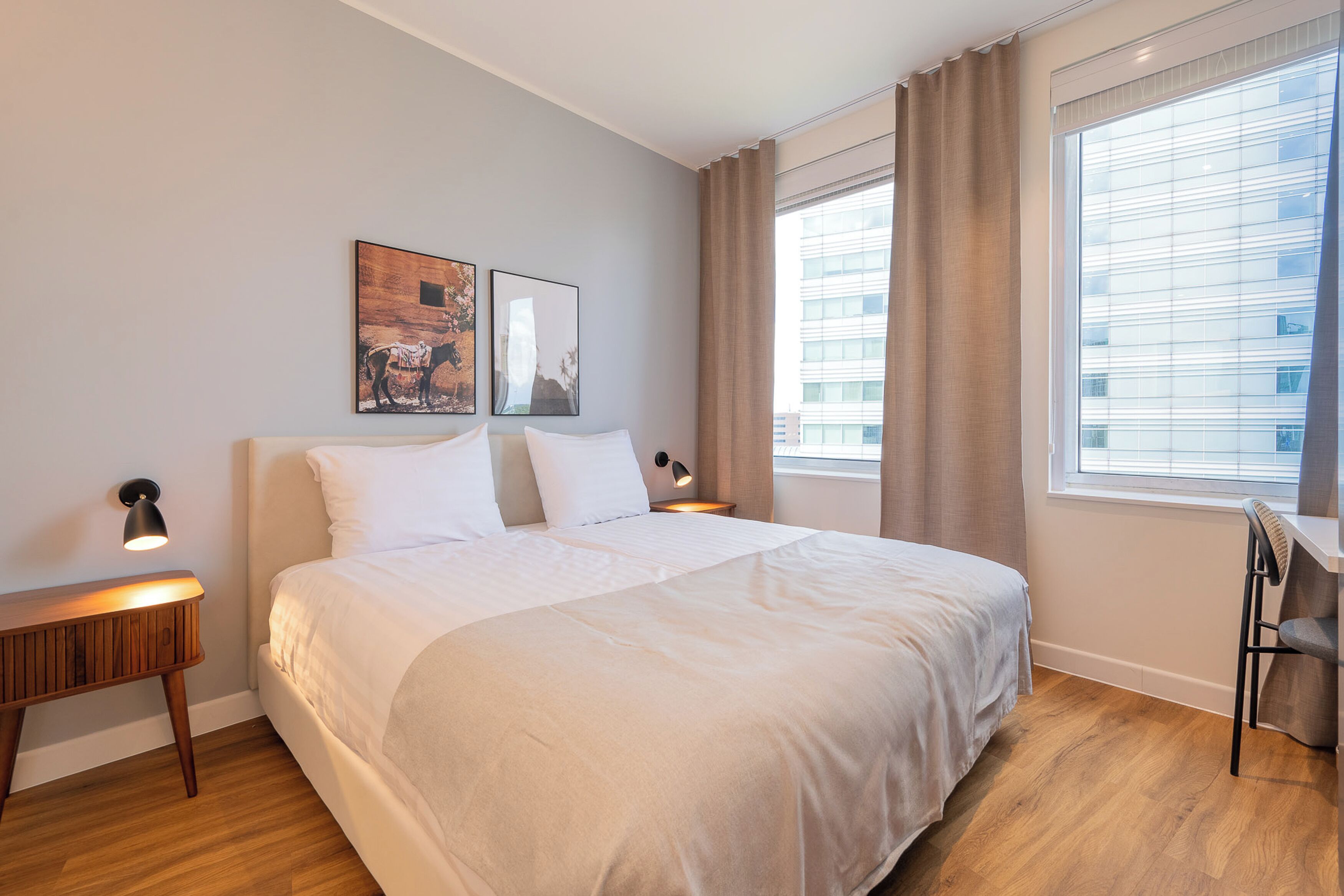 A modern hotel room with a large bed, a bedside table with a lamp, a small desk with a chair, and a large window with curtains.