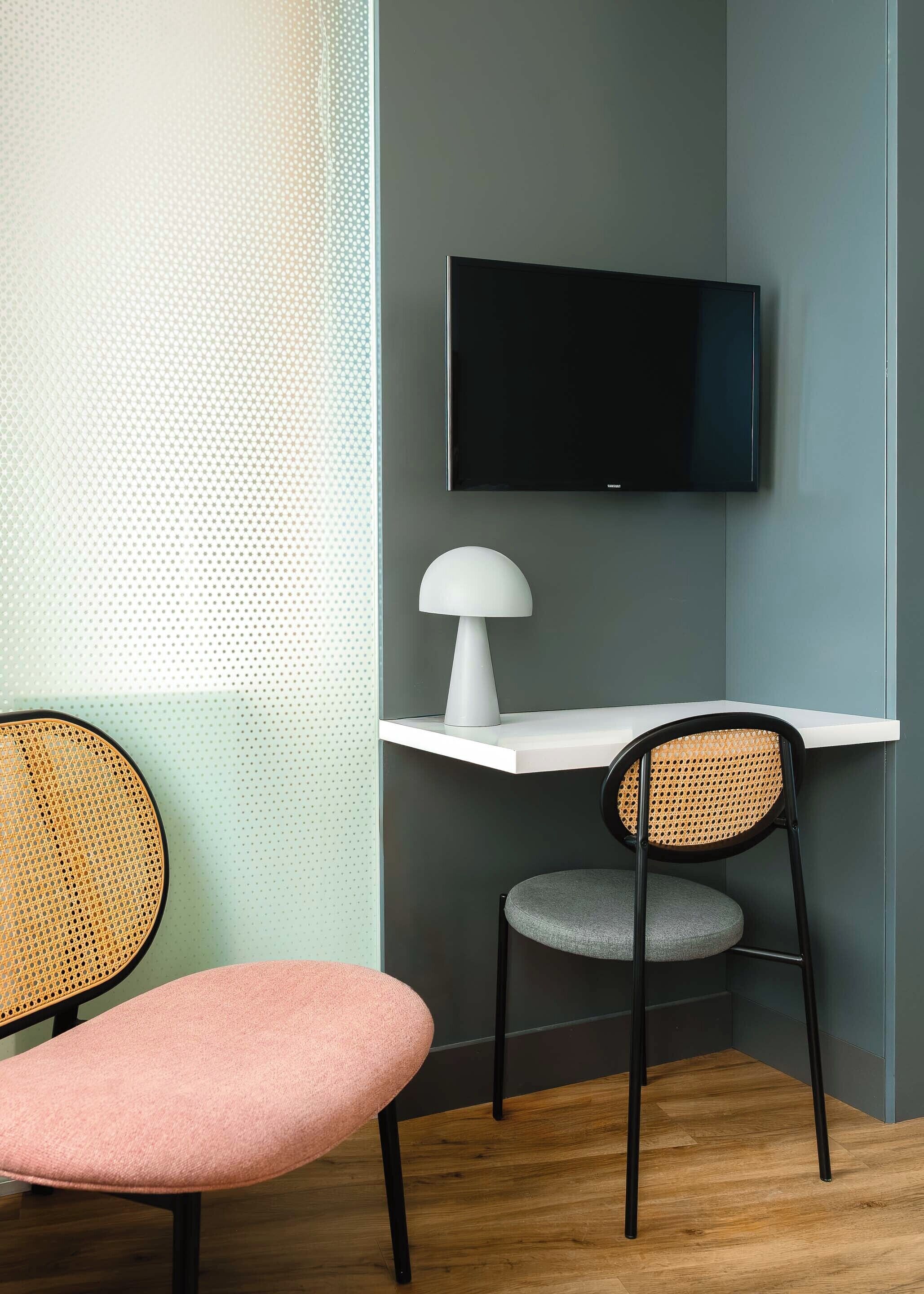 A corner room with a flat-screen TV mounted on the wall, a white desk, a chair with a woven backrest, and a pink cushioned bench.