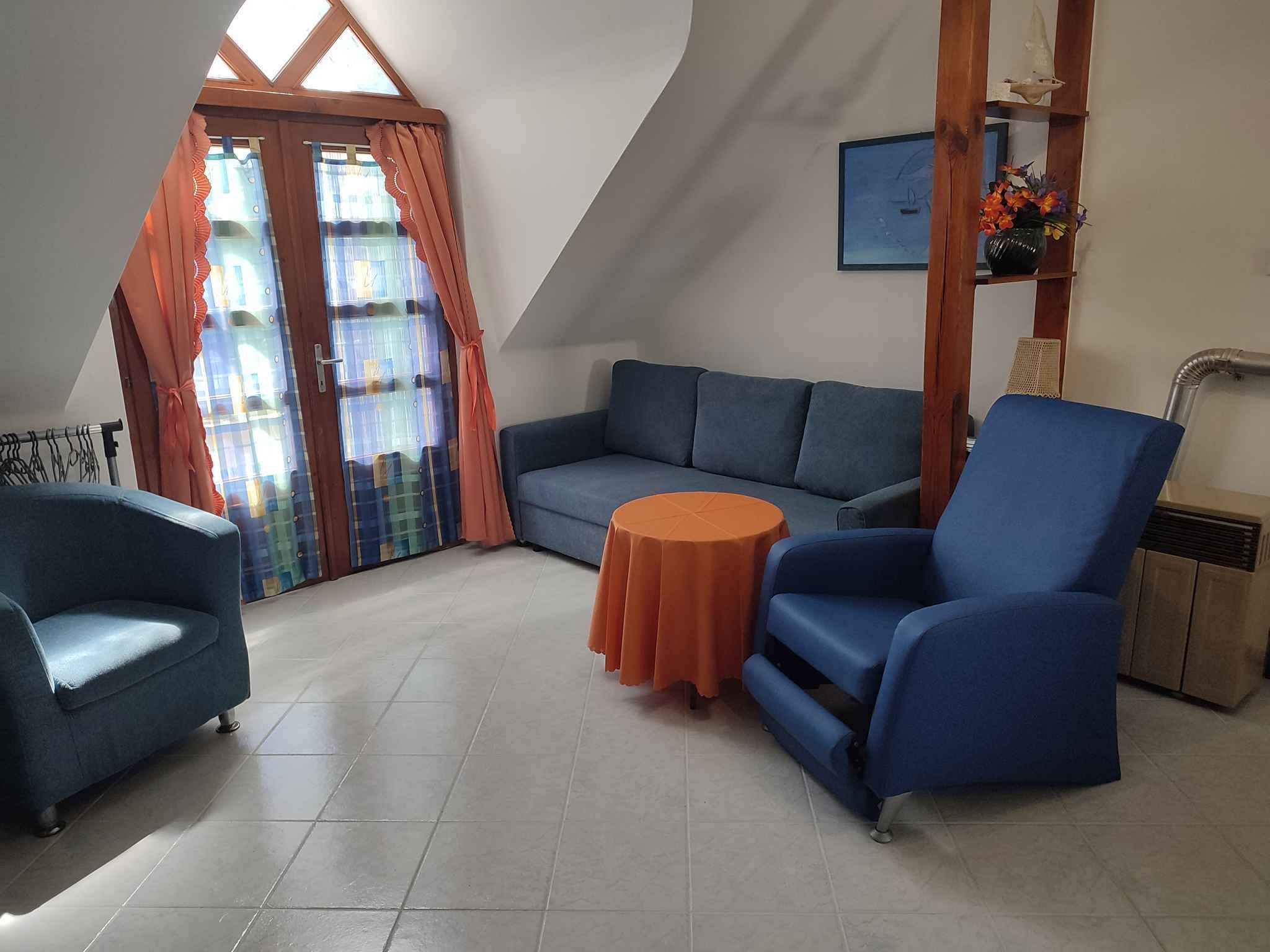 A room with a blue sofa, a blue armchair, and a small orange table. There is a window with curtains, a picture on the wall, and a shelf with decorative items.