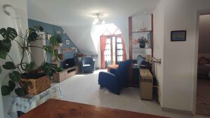 Amplio loft con techo alto, un ventanal con cortinas, un sofá azul, una mesa de madera, y una variedad de plantas y adornos.
