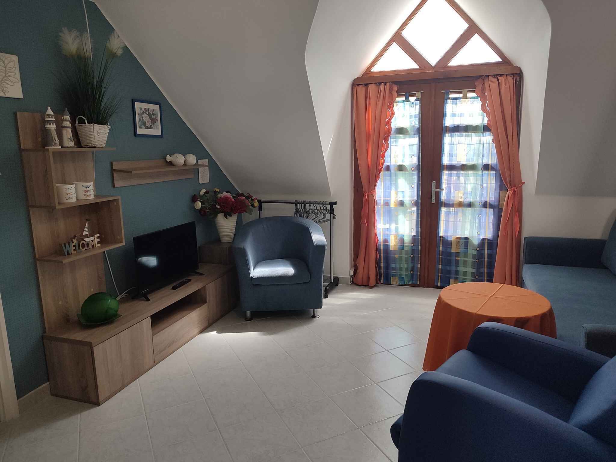 Una sala de estar acogedora con televisión, un sillón azul, un sofá y una mesita. Se observan estanterías con objetos decorativos y una ventana con cortinas.