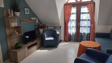 Una sala de estar acogedora con televisión, un sillón azul, un sofá y una mesita. Se observan estanterías con objetos decorativos y una ventana con cortinas.