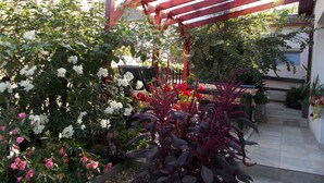 Un jardín con un porche de madera, diversas plantas en flor y una zona de patio con baldosas.