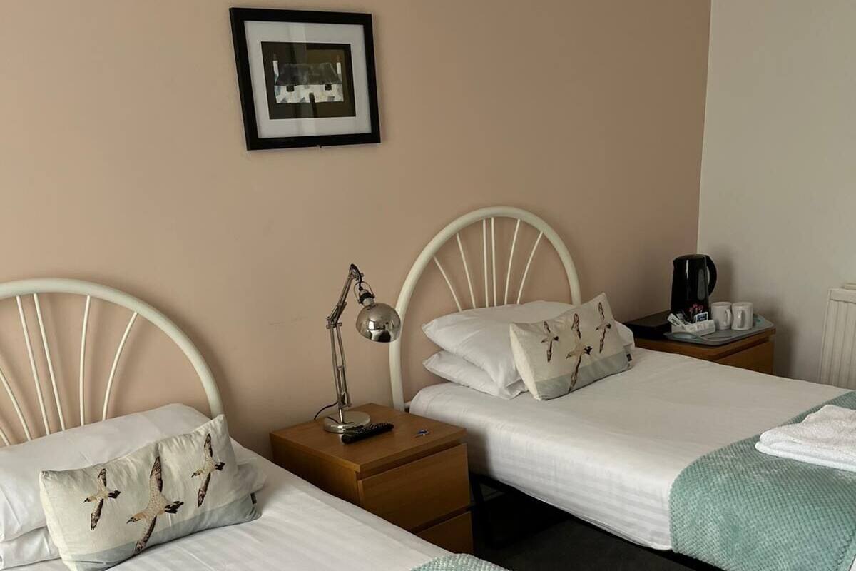 A hotel room with two beds, each with a white headboard, a bedside table with a lamp, and a framed picture on the wall.