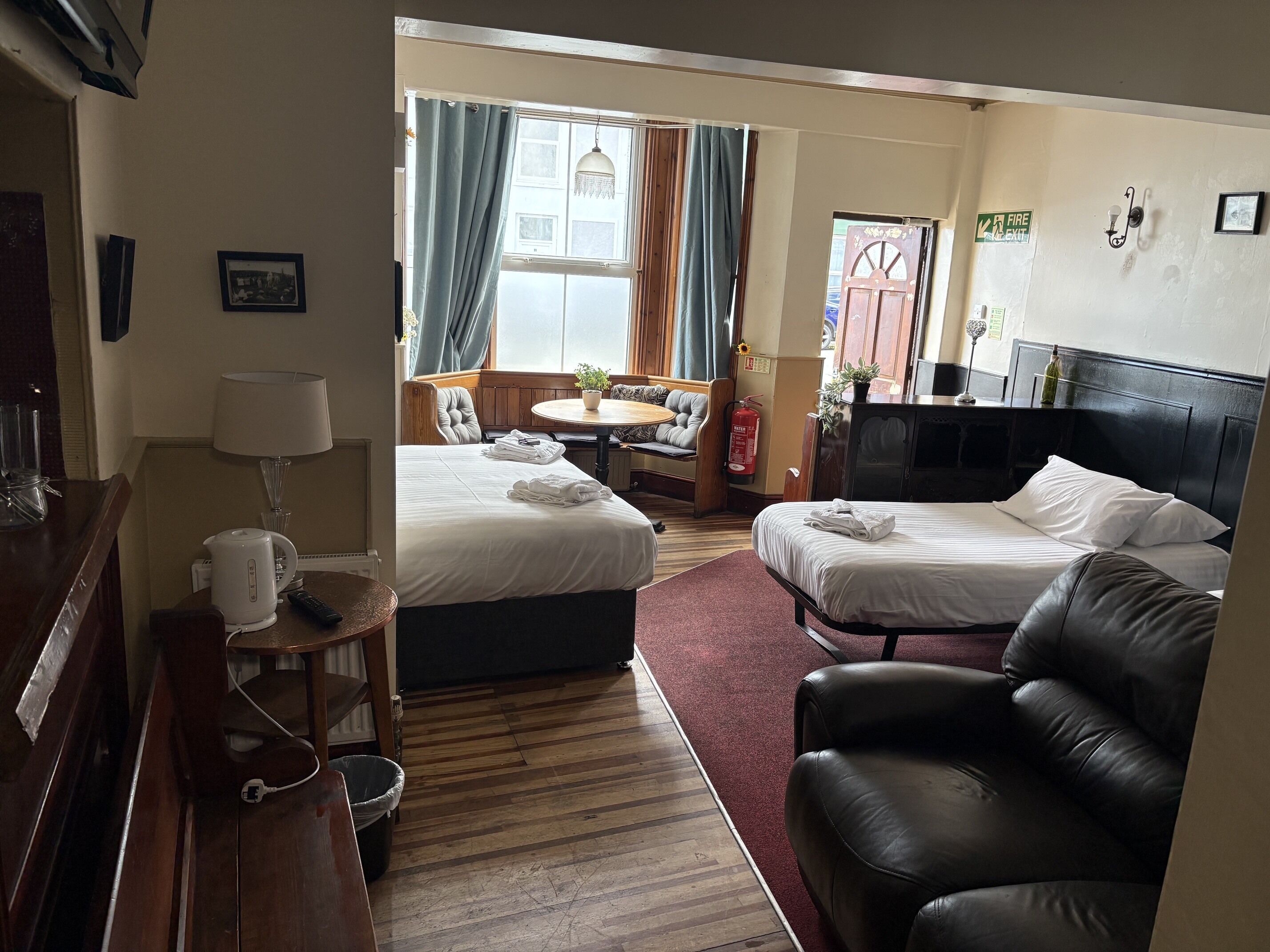 A hotel room with two beds, a sofa, a dining table, a lamp, and a fire extinguisher.