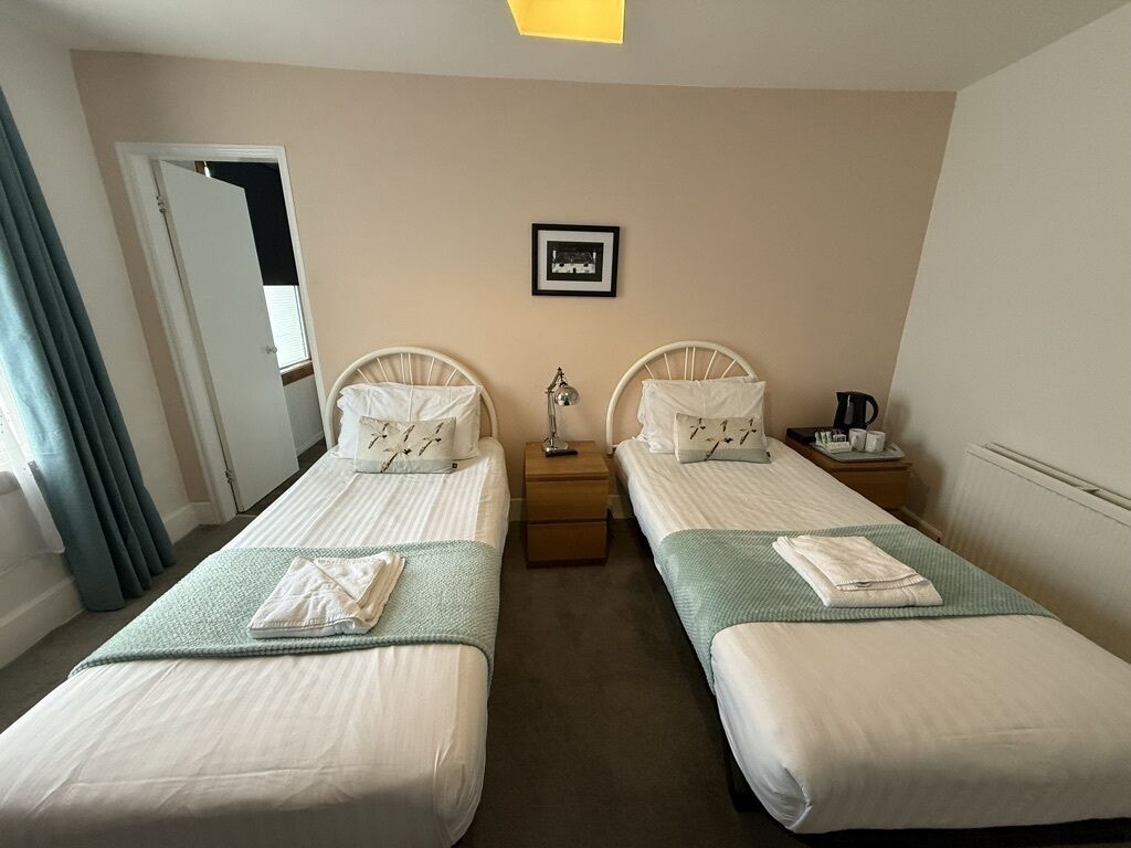 A hotel room with two single beds, a bedside table with a lamp, a framed picture on the wall, and a window with curtains.