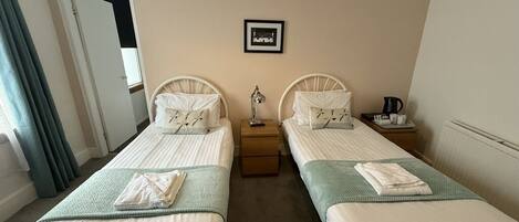 A hotel room with two single beds, a bedside table with a lamp, a framed picture on the wall, and a window with curtains.