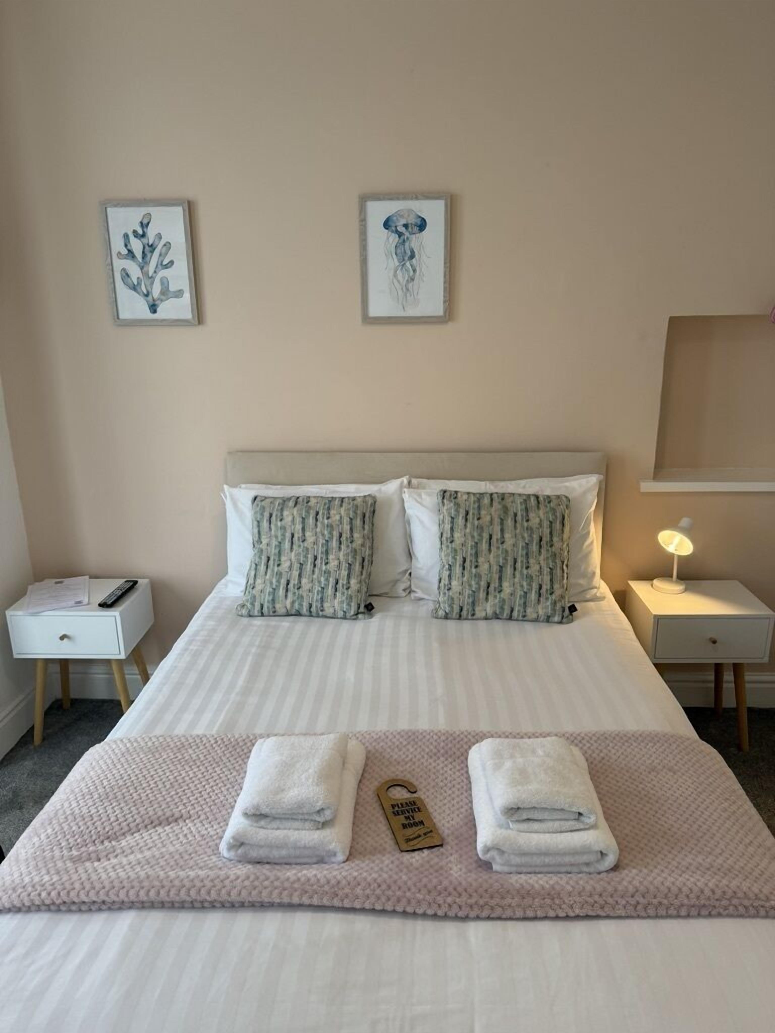 Un lit bien fait, avec une literie à rayures, deux oreillers, une table de chevet avec une lampe, et deux œuvres d’art encadrées accrochées au mur.
