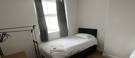 Una habitación pequeña con cama individual, piso de madera, ventana con cortinas y un perchero con ropa.
