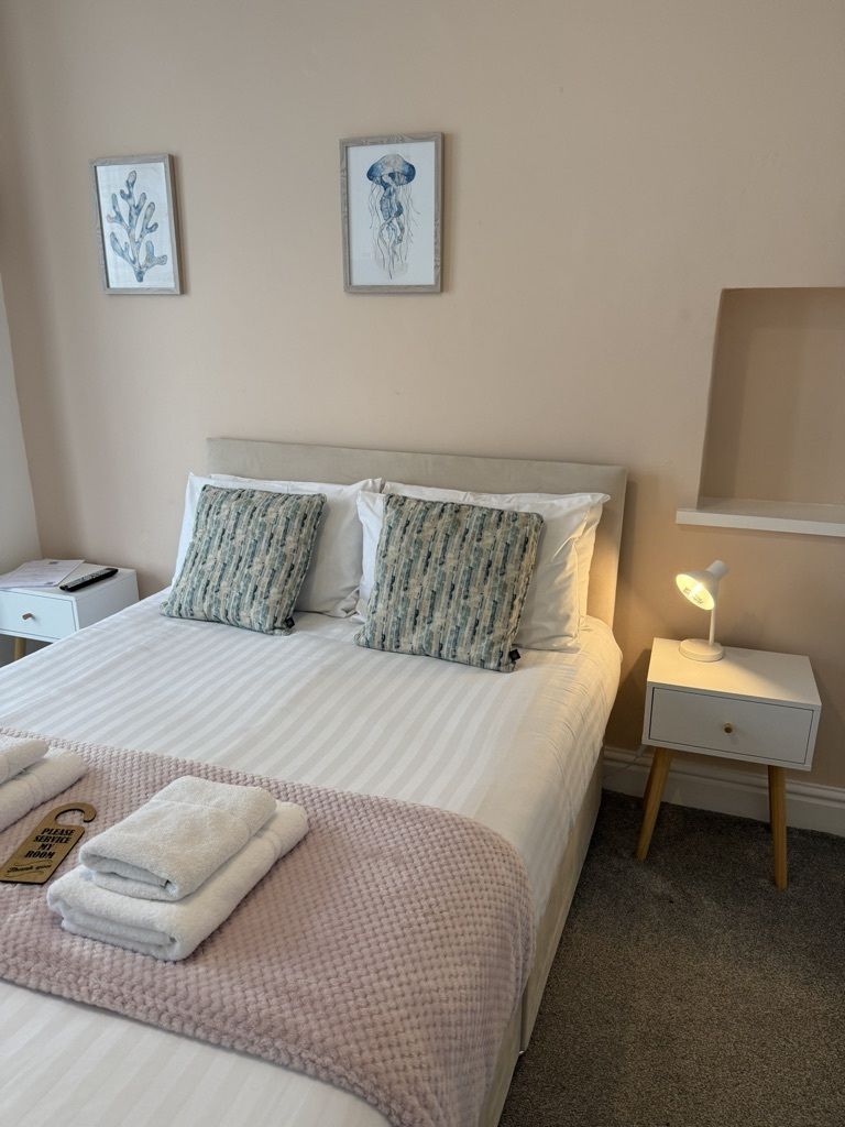 Ein ordentlich bezogenes Bett mit einer rosafarbenen Decke, zwei Kissen und einem gefalteten Handtuch auf der Seite. Ein Nachttisch mit einer Lampe und einer kleinen Schublade steht daneben. Zwei gerahmte Bilder hängen an der Wand über dem Bett.