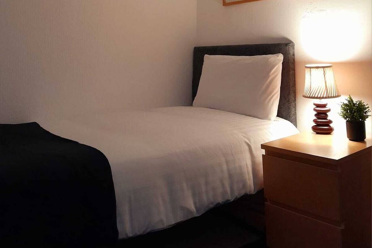 A bedroom with a bed, a nightstand with a lamp, a framed picture on the wall, and a ceiling light.