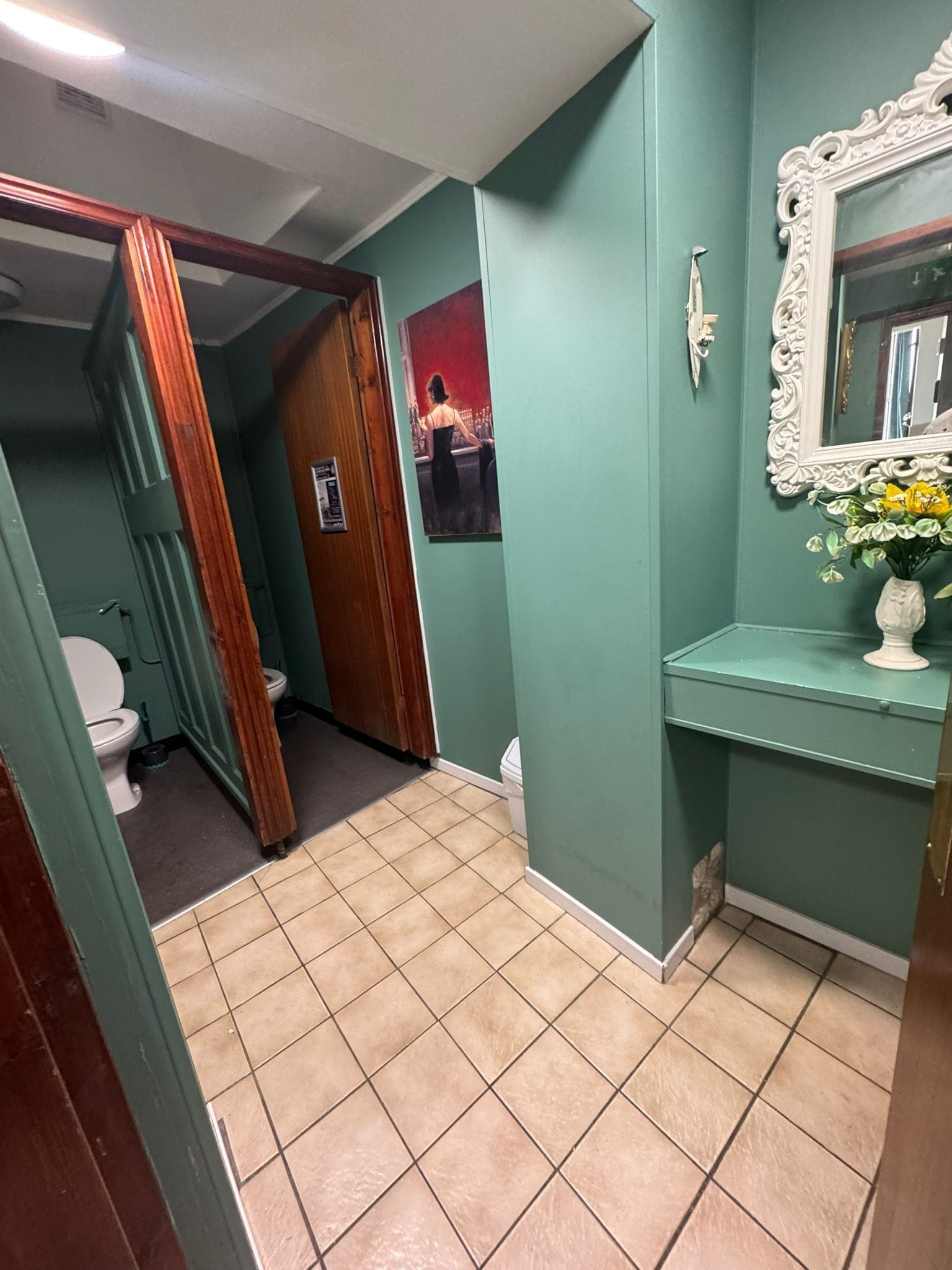 A bathroom with a toilet, a sink with a vase of flowers, a mirror with an ornate frame, and a poster on the wall.