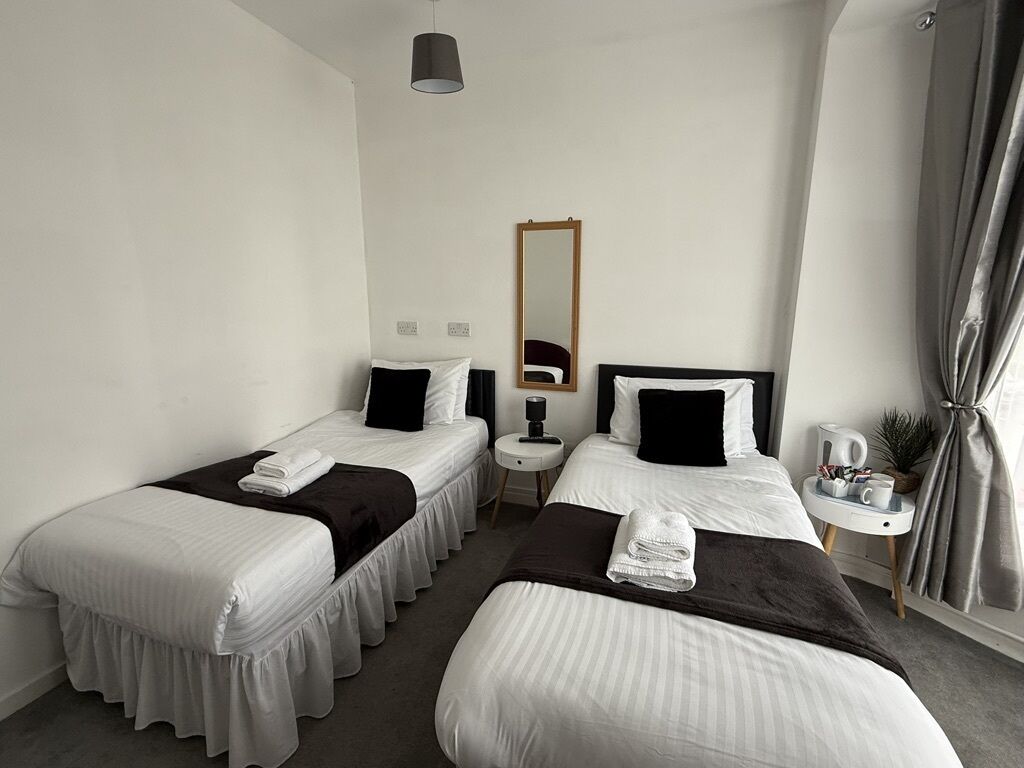 A hotel room with two single beds, a small bedside table with a lamp, a mirror, and a window with curtains.